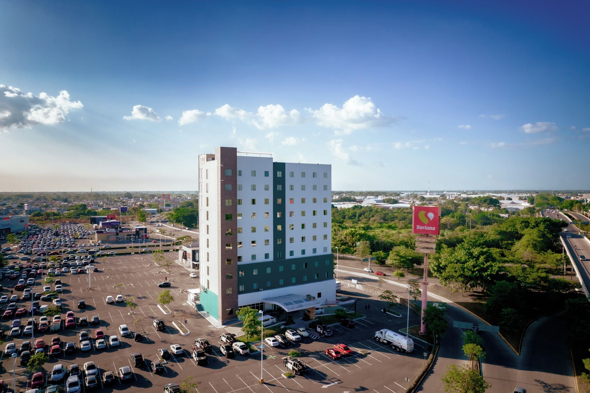 Un hotel de varios pisos con estacionamiento al frente, rodeado de vegetación y un cielo despejado.