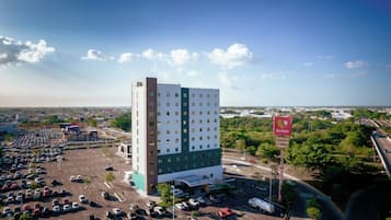 Un hotel de varios pisos con estacionamiento al frente, rodeado de vegetación y un cielo despejado.