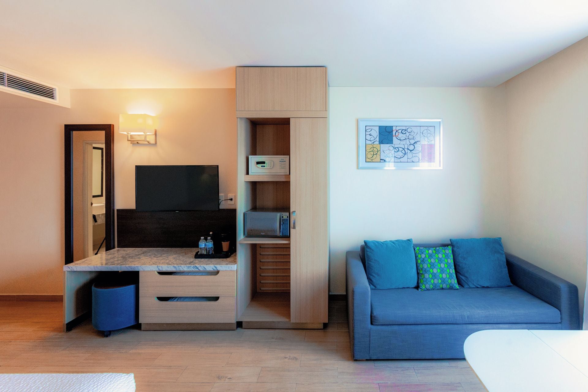 A compact living space with a kitchenette, a flat-screen TV, a sofa with patterned cushions, and a framed abstract art piece on the wall.