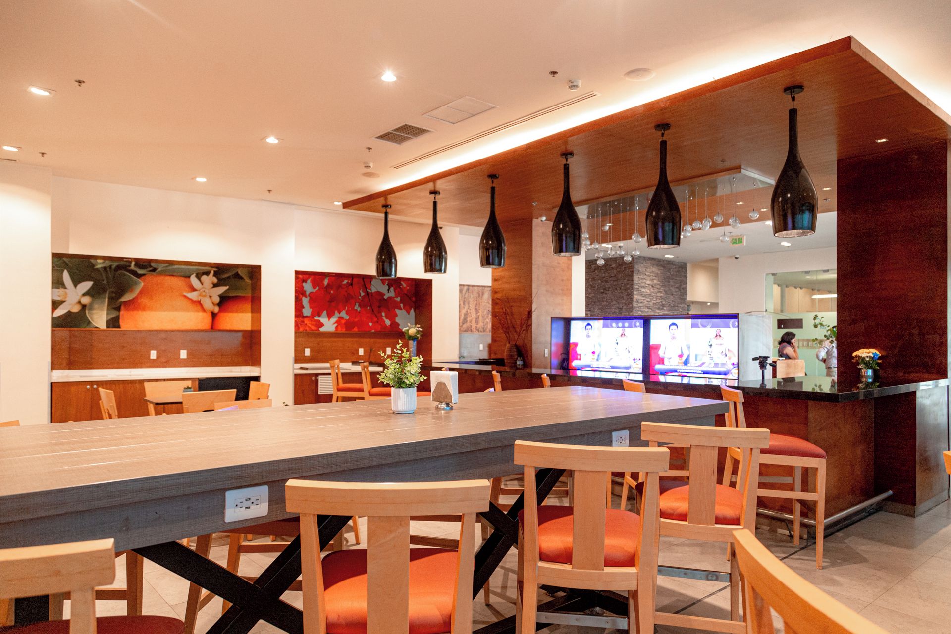 Un restaurante con una larga mesa compartida, sillas de madera, lámparas colgantes y una pantalla de televisión que muestra un programa.