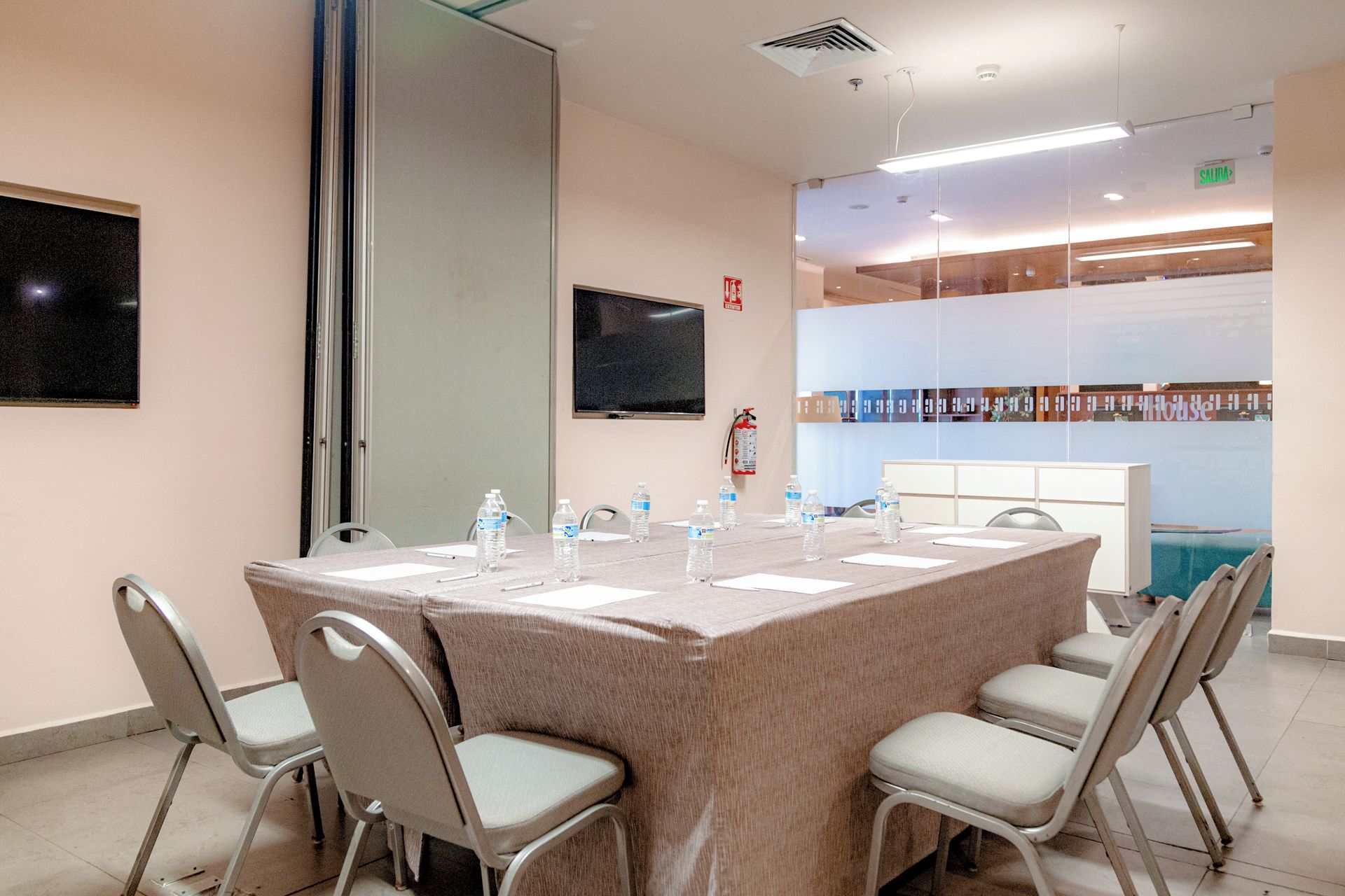A conference room with a large table, chairs, water bottles, a fire extinguisher, and a television on the wall.