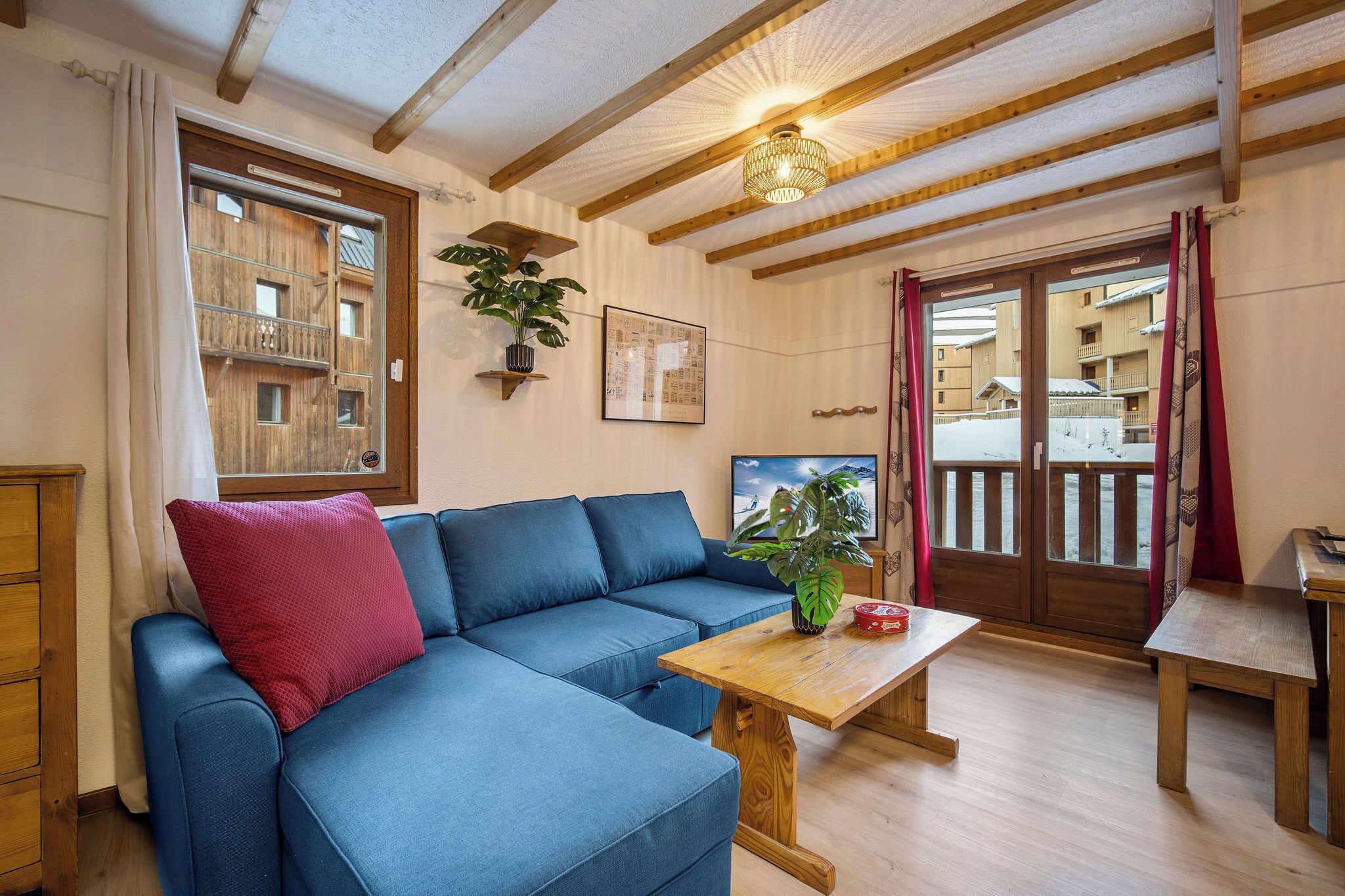 A cozy living room with a blue sofa, wooden ceiling beams, a wooden table, and a balcony view.