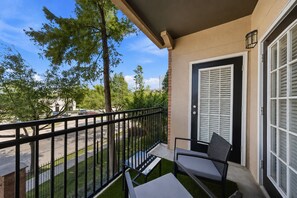 A balcony with a chair and table, a door with blinds, and a view of trees and a building in the background. - Modern 2BR 2BA Near NRG Stadium & Museum District (Houston)