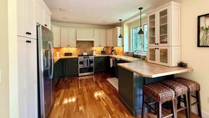 A kitchen with wooden flooring, white cabinets, a stainless steel refrigerator, a stove, and a sink. There are three bar stools and a window with a view of trees. - Cherry Bear Cottage at Woodstone in Glen Arbor (Glen Arbor)