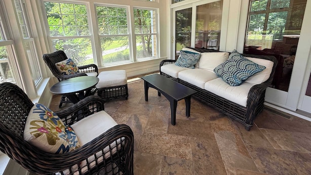 A sunroom with wicker furniture, a sofa with patterned cushions, and large windows offering a view of greenery. - Cherry Bear Cottage at Woodstone in Glen Arbor (Glen Arbor)