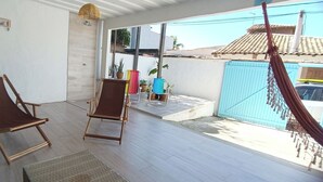 Uma área externa coberta com duas cadeiras de madeira, uma rede e vista para um bairro residencial.
