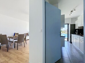 Eine moderne Küche mit weißen Schränken und einer schwarzen Arbeitsplatte, ein Essbereich mit einem Holztisch und Stühlen sowie Blick auf einen Swimmingpool durch eine offene Tür.