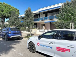 Une voiture blanche, portant le logo « interhome », est garée devant un immeuble doté de balcons et d’un toit bleu.
