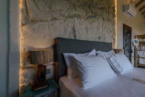 A bedroom with a stone wall, a bed with white bedding, a wooden nightstand with a lamp, and a wooden shelf with bags. - AER - Romantic Private Pool Villa with Spectacular 180° Ocean Views (Korissia)