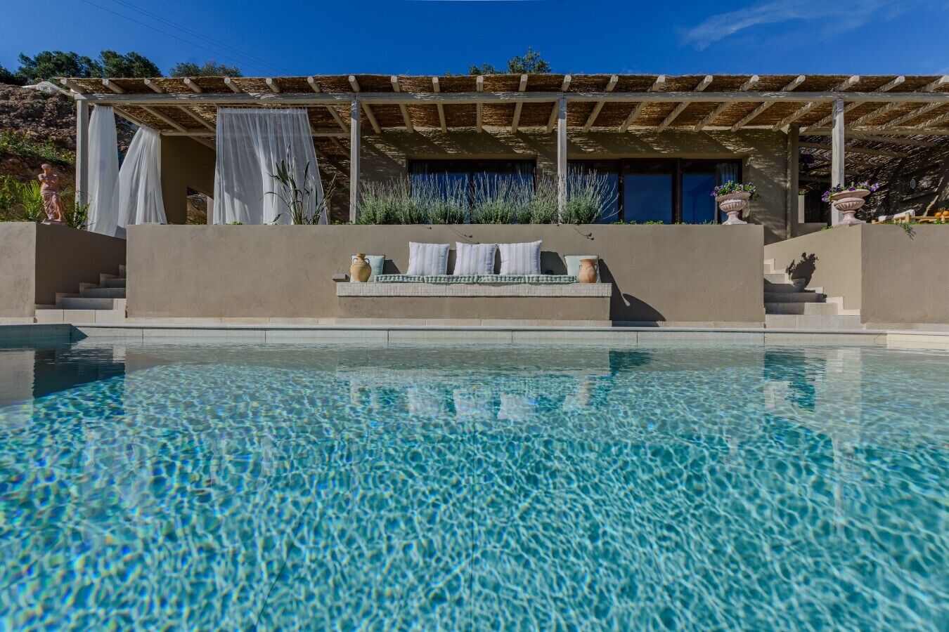 A swimming pool with a clear blue surface, a covered seating area with cushions, and a stone statue.