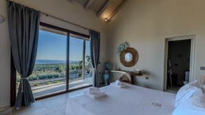 A bedroom with a large bed, a view of the landscape, a vanity with a mirror, and a door leading to another room. - Romantic Private Pool Villa with Spectacular 180° Ocean Views (Korissia)