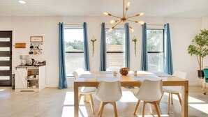 A dining area with a wooden table, white chairs, a chandelier, and large windows with curtains. - Hill Country Hideaway | Modern Barndominium for 6 (Dripping Springs)