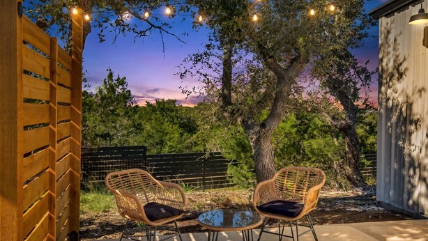 An outdoor seating area with two wicker chairs and a glass table, surrounded by trees and a wooden fence. - Hill Country Hideaway | Modern Barndominium for 6 (Dripping Springs)