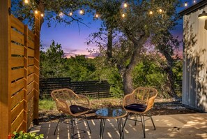 An outdoor seating area with two wicker chairs and a glass table, surrounded by trees and a wooden fence. - Hill Country Hideaway | Modern Barndominium for 6 (Dripping Springs)