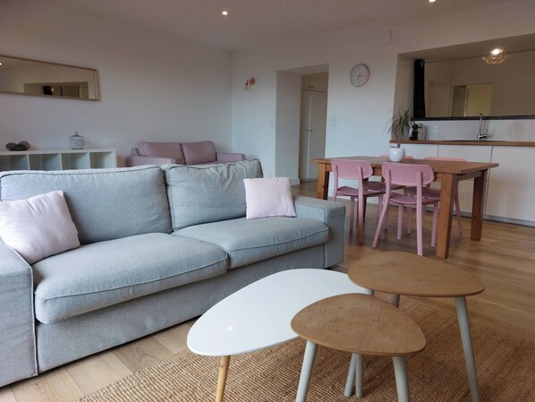 A modern living room with a grey sofa, a white coffee table, and a wooden dining table with pink chairs.