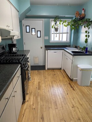 Una cocina con suelo de madera, armarios blancos, estufa, lavavajillas y una ventana con persianas.