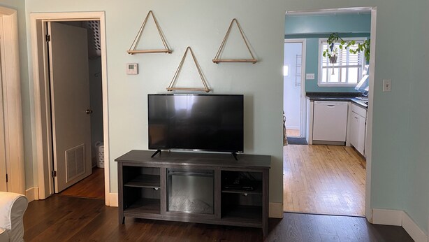A living room with a flat-screen TV on a dark wooden entertainment center, a door leading to a hallway, and a view into a kitchen area. - Perfect 1-bedroom house in welcoming Ontonagon for a relaxing stay (Ontonagon)