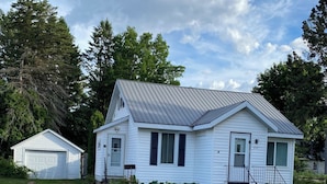 A single-story white house with a metal roof, a front porch, and a driveway leading to a garage. - Perfect 1-bedroom house in welcoming Ontonagon for a relaxing stay (Ontonagon)