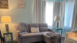 A living room with a grey sectional sofa, a small table with a lamp, a chair, and a window with curtains. - Perfect 1-bedroom house in welcoming Ontonagon for a relaxing stay (Ontonagon)