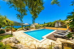 A swimming pool with lounge chairs and umbrellas, surrounded by stone flooring and greenery. - Villa Apanemia - Three Bedroom Villa, Sleeps 6 (Kassiopi)