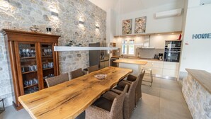 A modern kitchen with a stone wall, wooden dining table, wicker chairs, and a built-in cabinet with glassware. - Villa Apanemia - Three Bedroom Villa, Sleeps 6 (Kassiopi)
