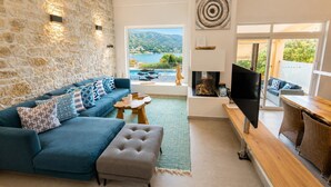 A living room with a stone wall, a sofa with patterned cushions, a television, and a view of a body of water. - Villa Apanemia - Three Bedroom Villa, Sleeps 6 (Kassiopi)