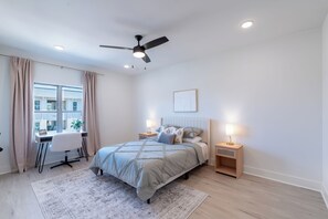 Ein Schlafzimmer mit Bett, Nachttischen, Schreibtisch, Stuhl, Deckenventilator und einem Fenster mit Vorhängen.
