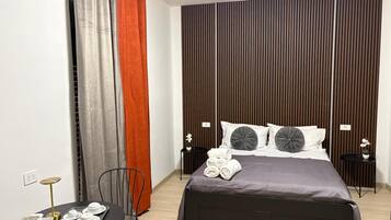 Una camera d'albergo moderna con un letto, un tavolino con una sedia, un comodino e una parete con persiane verticali.