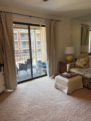 A living room with a sliding glass door leading to a balcony, a patterned sofa, a small ottoman, and a lamp. - Cozy Beach Escape (Ocean City)