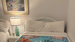 A bed with a beach-themed bedspread, a nightstand with a lamp and a decorative item, and a framed picture on the wall. - Cozy Beach Escape (Ocean City)