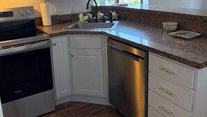 A compact kitchen with white cabinets, a stainless steel dishwasher, a built-in oven, and a window providing a view of the outside. - Cozy Beach Escape (Ocean City)
