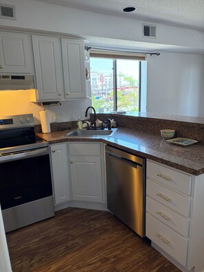 A compact kitchen with white cabinets, a stainless steel dishwasher, a built-in oven, and a window providing a view of the outside. - Cozy Beach Escape (Ocean City)