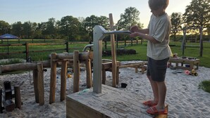 Een kind speelt op een houten constructie in een zandrijk buitengebied met een hek en begroeiing op de achtergrond.