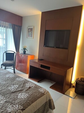 Ein Hotelzimmer mit einem Bett, einem Schreibtisch mit Fernseher, einem Sessel, einer Topfpflanze und einem gerahmten Bild an der Wand.