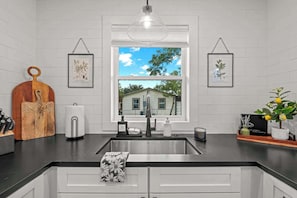 Cozinha com uma janela grande, armários brancos, bancada preta e luminária pendente.