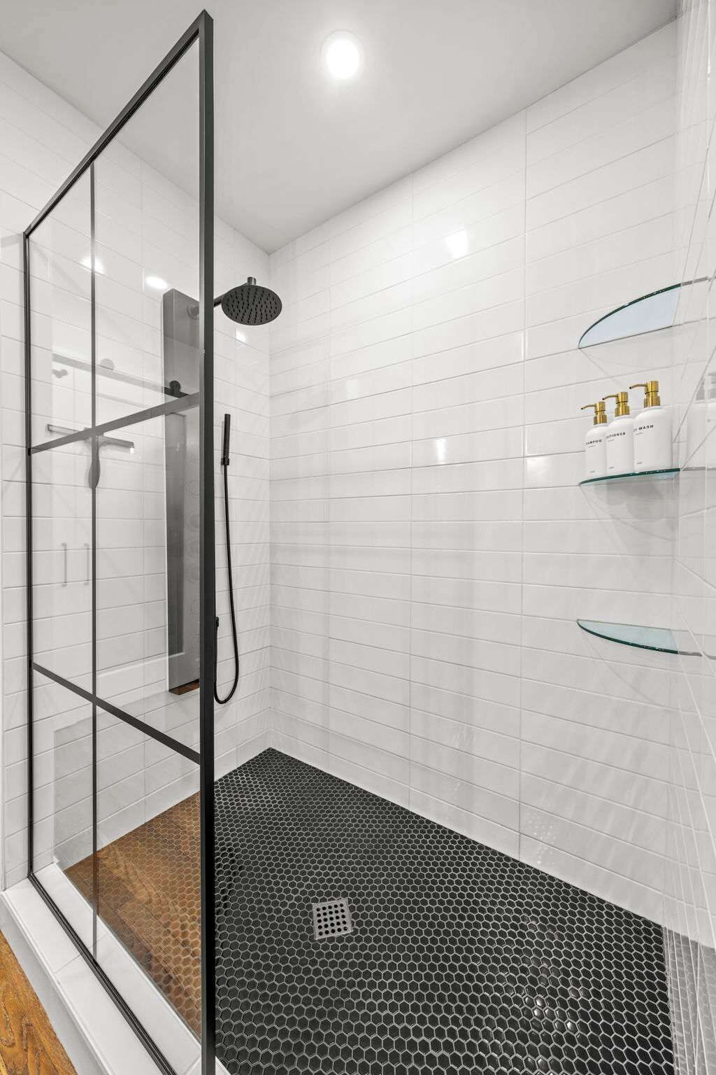 A modern bathroom with a glass shower enclosure, white tiled walls, and a black hexagonal floor.