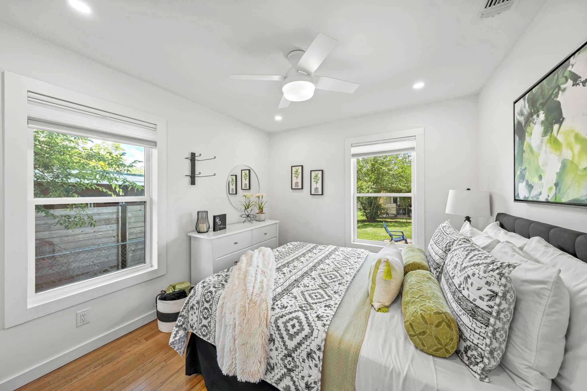 A bedroom with a bed, two windows, a ceiling fan, and a view of greenery outside.