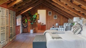 Quarto no sótão com teto de madeira, cama, uma mesinha, cadeira e planta em vaso.