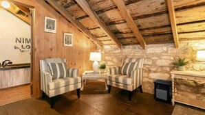 Um quarto rústico com vigas de madeira, paredes de pedra, duas poltronas xadrez, uma mesa pequena e um abajur.
