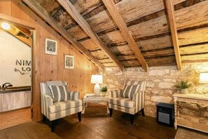 A rustic room with wooden beams, stone walls, two plaid armchairs, a small table, and a lamp. - Nimitz Loft - 1855 Historic Loft on Main St! (Fredericksburg)
