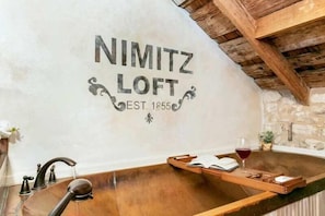 A rustic bathroom with a wooden bathtub, a vintage Nimitz Loft sign, and a stone wall. - Nimitz Loft - 1855 Historic Loft on Main St! (Fredericksburg)