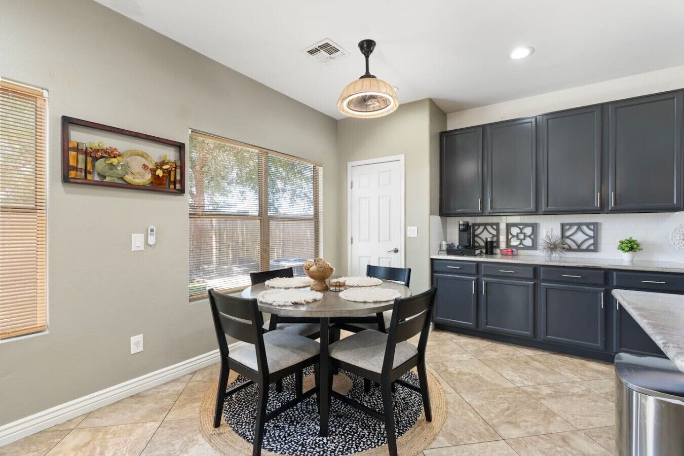A kitchen with dark cabinets, a round dining table with chairs, a pendant light, and a window with blinds.