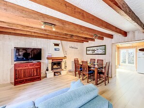 Uma sala de estar acolhedora com lareira, vigas de madeira no teto, uma mesa de jantar com cadeiras, um sofá e uma televisão.