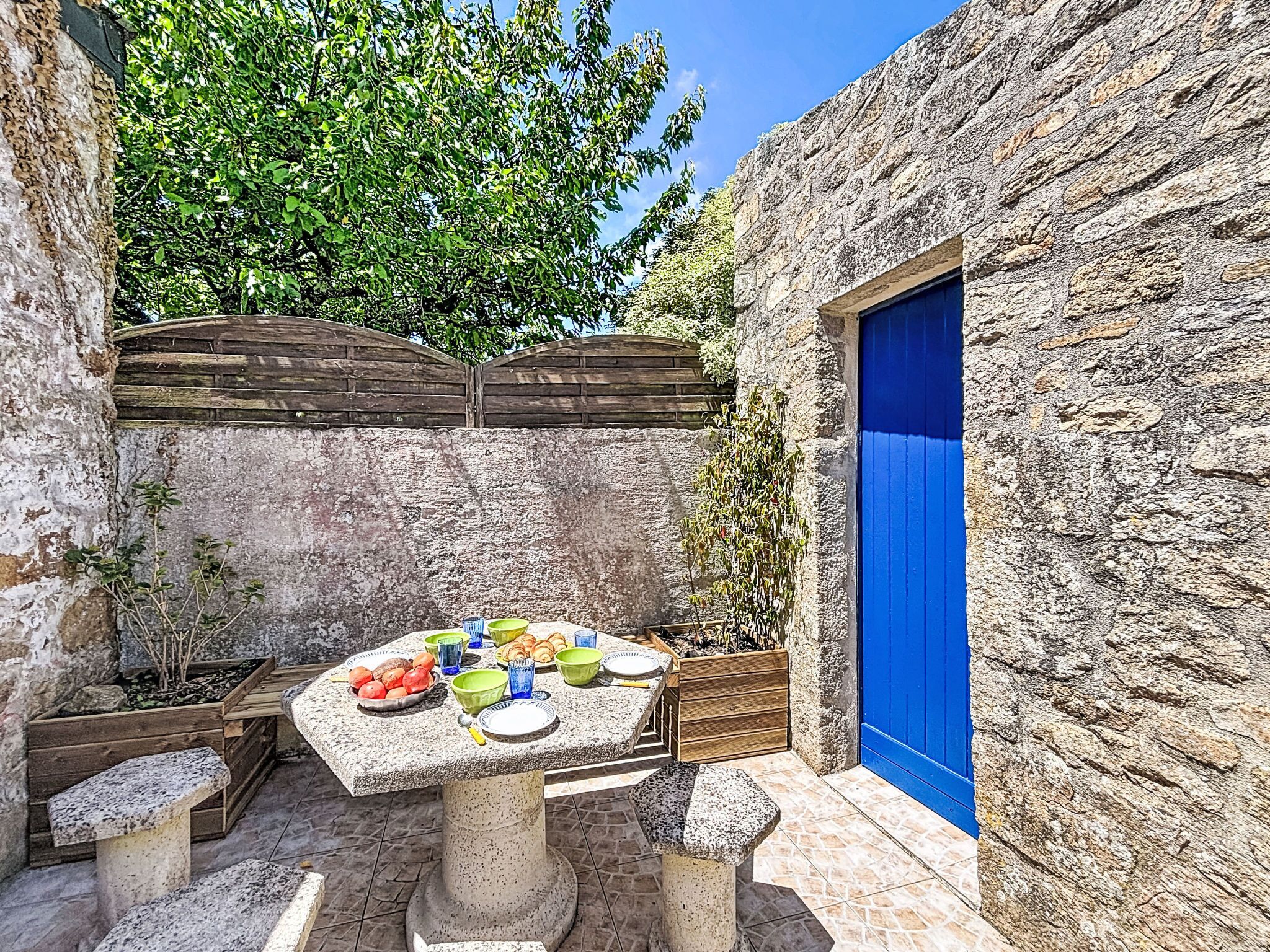 A stone wall with a blue door, a table with fruits and bowls, and a wooden fence.