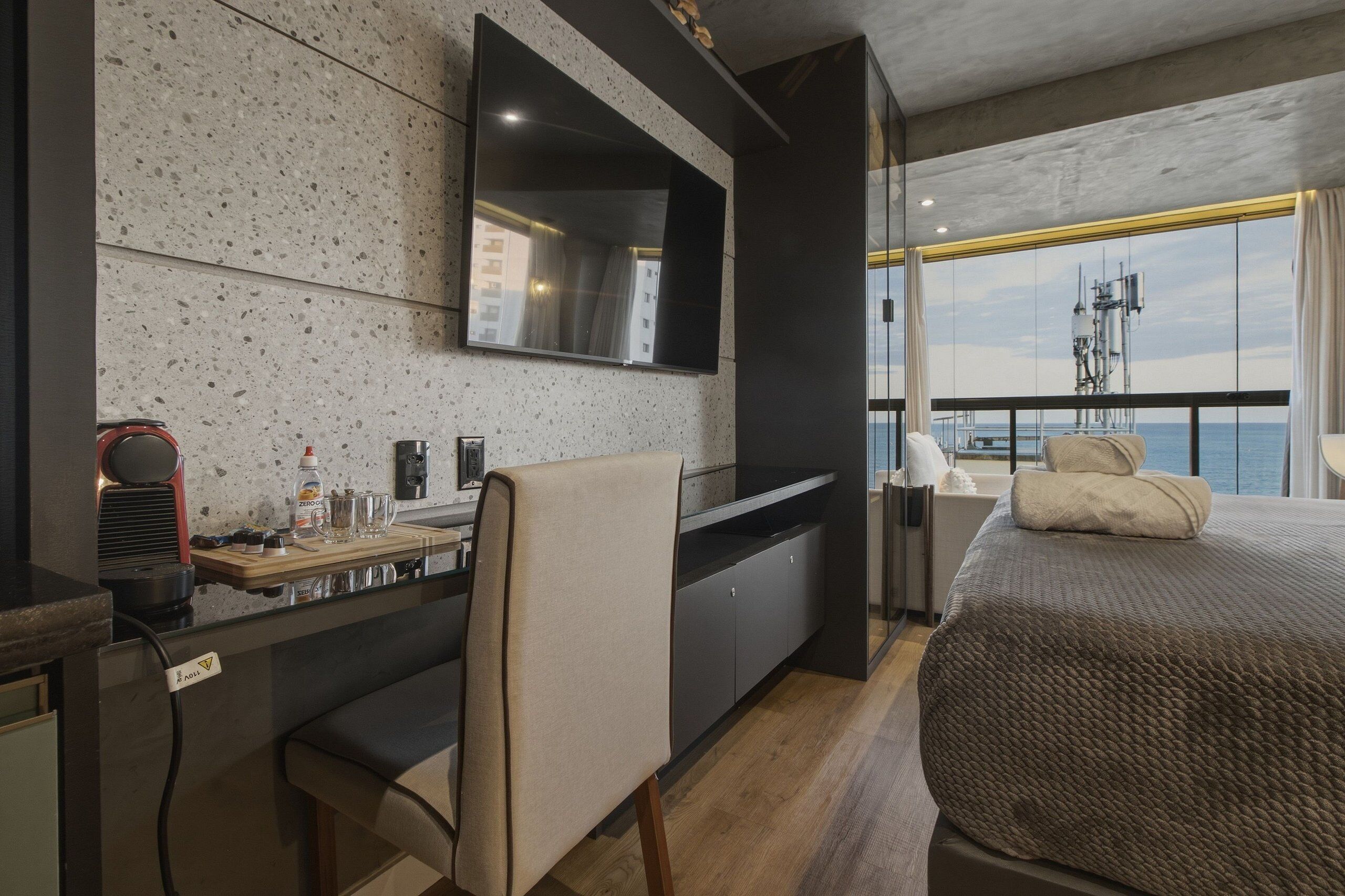 A modern hotel room with a large bed, a desk with a chair, a flat-screen TV mounted on the wall, and a balcony with a view of the sea.
