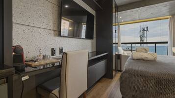 A modern hotel room with a large bed, a desk with a chair, a flat-screen TV mounted on the wall, and a balcony with a view of the sea.