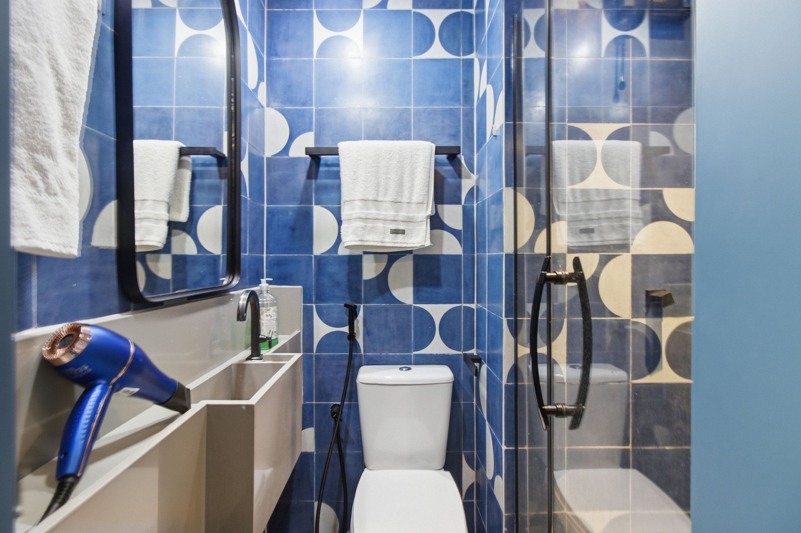 Une salle de bain avec des carreaux bleus à motifs, des toilettes blanches, un porte-serviettes, un miroir et un sèche-cheveux posé sur une étagère.