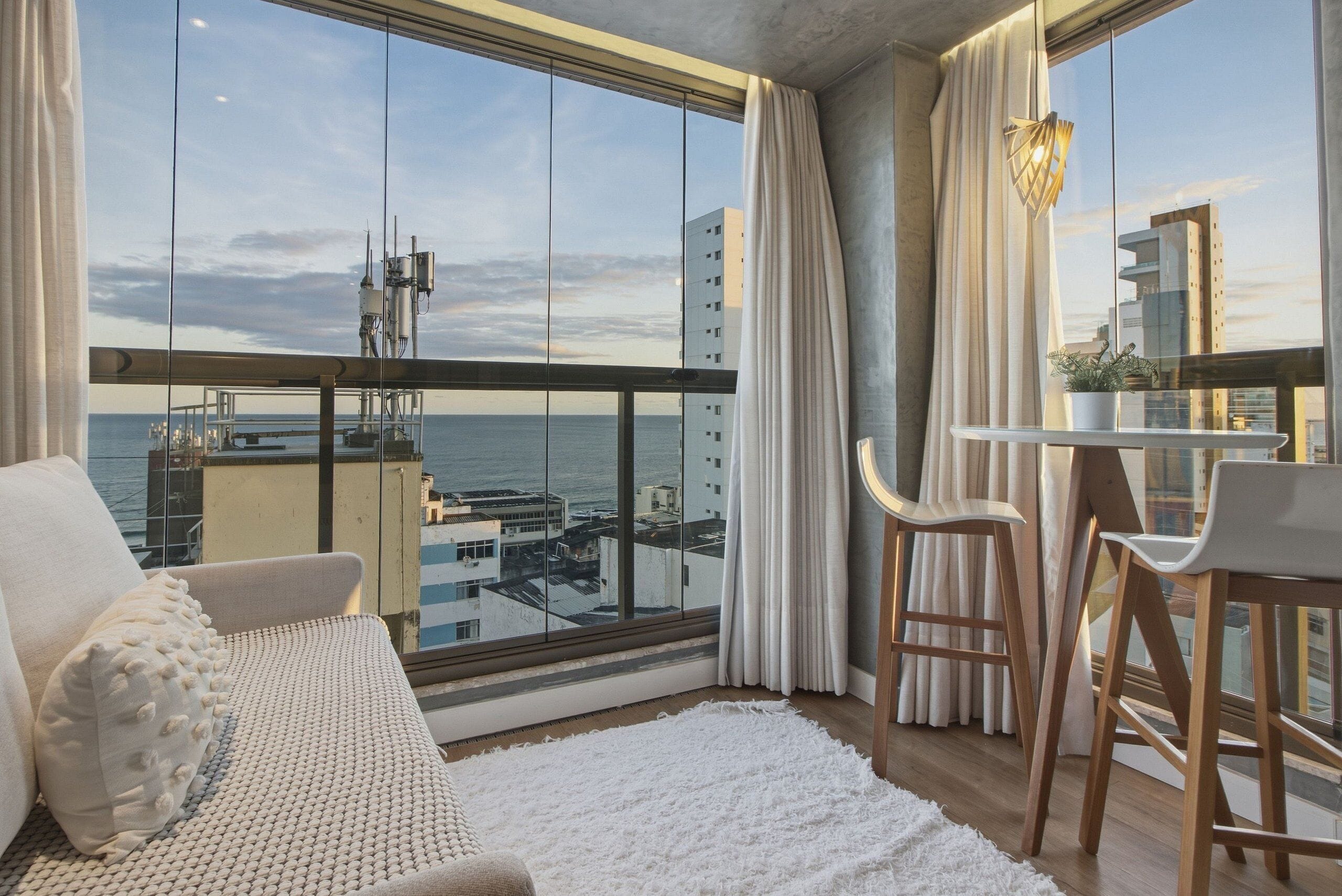 A balcony with a view of the sea and buildings, a sofa, a small table with chairs, and a white rug.