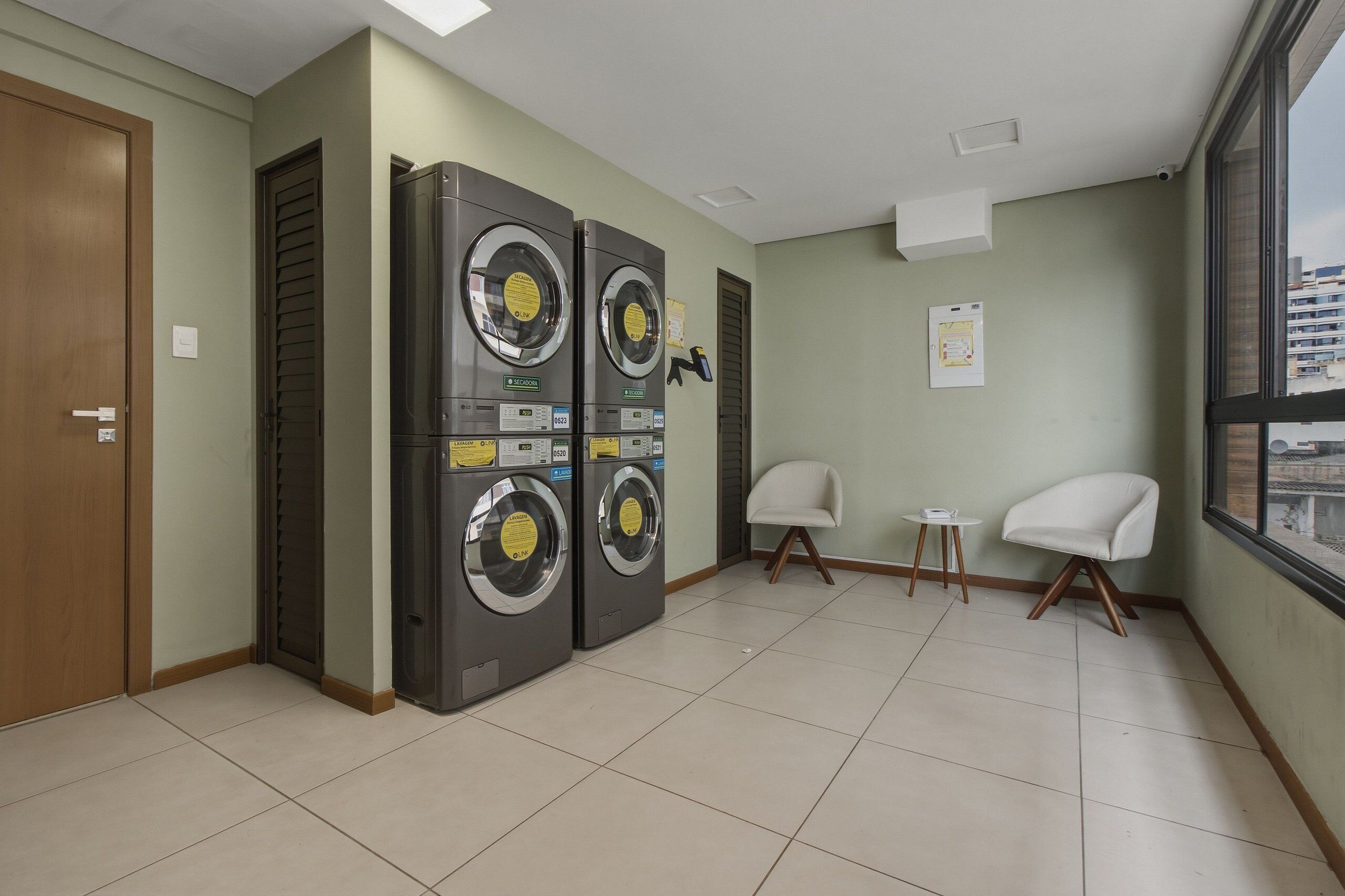 A laundry room with four washing machines, two chairs, a small table, and a window with a view of buildings.