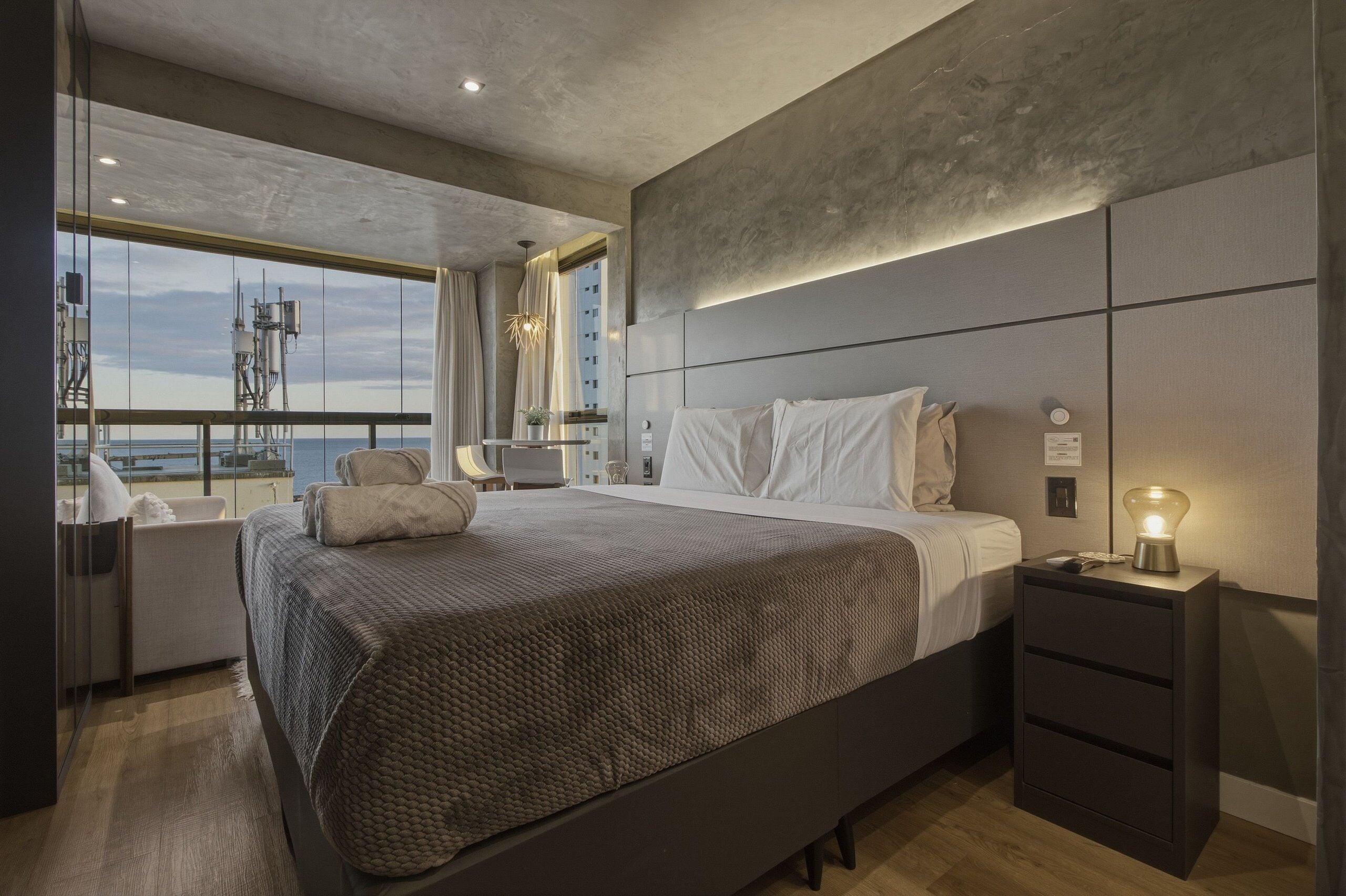 Quarto moderno com uma cama grande, mesas de cabeceira e um criado-mudo com abajur, além de vista para a cidade através de amplas janelas.